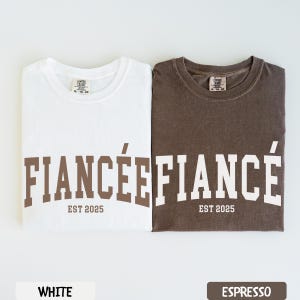 K&ouml;nnte beinhalten: Zwei gefaltete T-Shirts, eines wei&szlig; und eines espressofarben, mit den W&ouml;rtern "FIANC&Eacute;E" und "FIANC&Eacute;" in Braun bzw. Wei&szlig; bedruckt. Der Text "EST 2025" steht darunter. Das wei&szlig;e Shirt hat ein wei&szlig;es Etikett mit der Aufschrift "WHITE" und das Espresso-Shirt ein Etikett mit der Aufschrift "ESPRESSO".