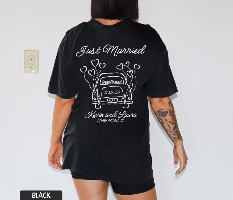 May include: Black t-shirt with a white graphic design on the back. The design features the words "Just Married" above a car illustration with heart-shaped balloons. Below the car are the names "Kevin and Laura" and the location "Charleston, SC".