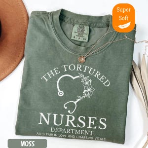 May include: A moss green t-shirt with the text "THE TORTURED NURSES DEPARTMENT" in white. The design includes a heart-shaped stethoscope and floral accents. The shirt is labeled "Super Soft" and is a Comfort Colors brand.