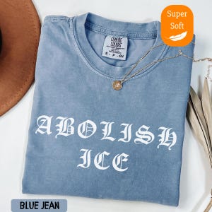 May include: A blue jean-colored t-shirt with the words "ABOLISH ICE" in white gothic lettering. The shirt has a crew neck and short sleeves. A gold necklace with a pendant rests on the shirt. The shirt is labeled "Super Soft."