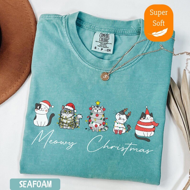 May include: Seafoam green t-shirt with a "Meowy Christmas" graphic featuring five cartoon cats in festive attire. The cats are decorated with Santa hats, Christmas lights, and scarves. The shirt has a "Super Soft" label.