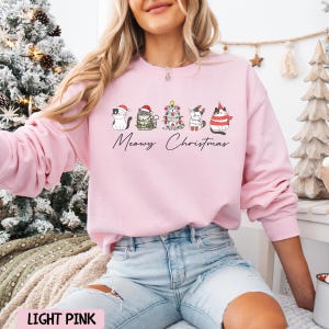 May include: Light pink sweatshirt with a graphic of five cats decorated for Christmas. The cats are wearing Santa hats, scarves, and a Christmas tree costume. The text "Meowy Christmas" is below the cats.