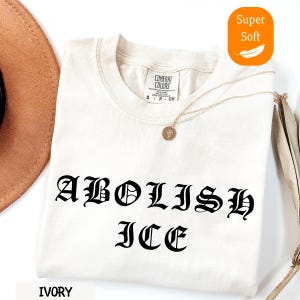May include: Ivory-colored t-shirt with the words "ABOLISH ICE" in a gothic font. The shirt has a crew neck and short sleeves. A gold necklace with a pendant is draped across the shirt. The shirt is labeled "Super Soft".