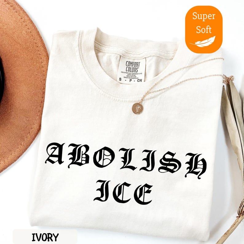 May include: Ivory-colored t-shirt with the words "ABOLISH ICE" in a gothic font. The shirt has a crew neck and short sleeves. A gold necklace with a pendant is draped across the shirt. The shirt is labeled "Super Soft".