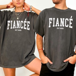 K&ouml;nnte beinhalten: Zwei anthrazitfarbene T-Shirts mit dem Wort "FIANC&Eacute;E" und "EST 2025" in Wei&szlig; aufgedruckt. Die Shirts haben einen Vintage-Look. Die T-Shirts sind kurz&auml;rmelig und haben eine lockere Passform.