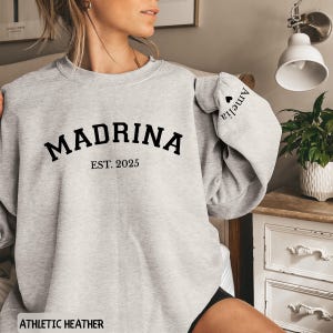 May include: Athletic heather gray sweatshirt with the word "MADRINA" in black block letters, with "EST. 2025" below. The sleeve has the name "Amelia" printed on it. The sweatshirt is a casual style.