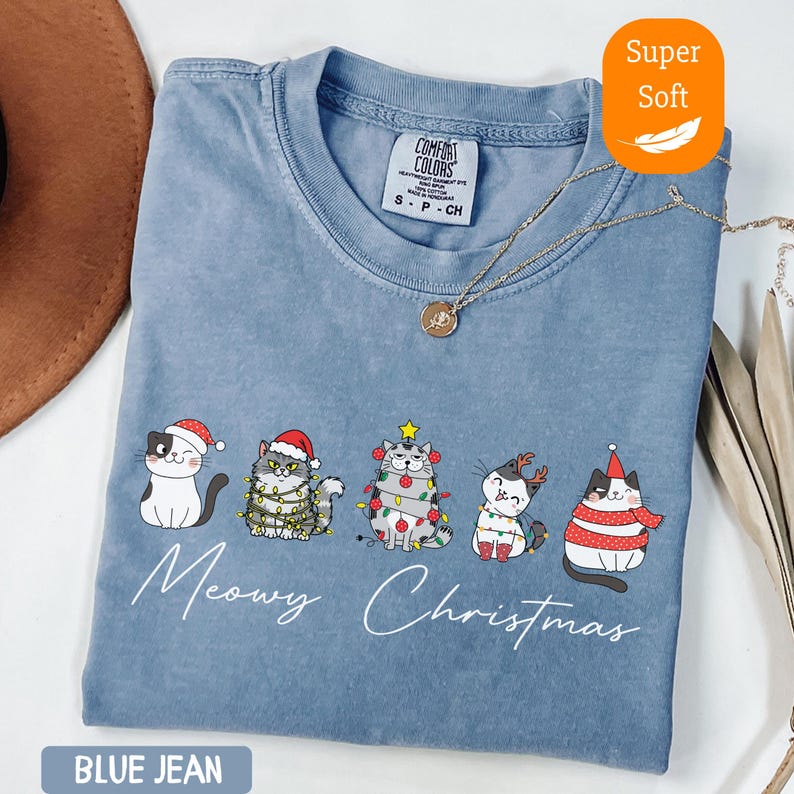 May include: Blue jean colored t-shirt with a "Meowy Christmas" graphic featuring five cartoon cats in festive holiday attire. The shirt has a crew neck and short sleeves. A gold necklace is draped across the shirt.