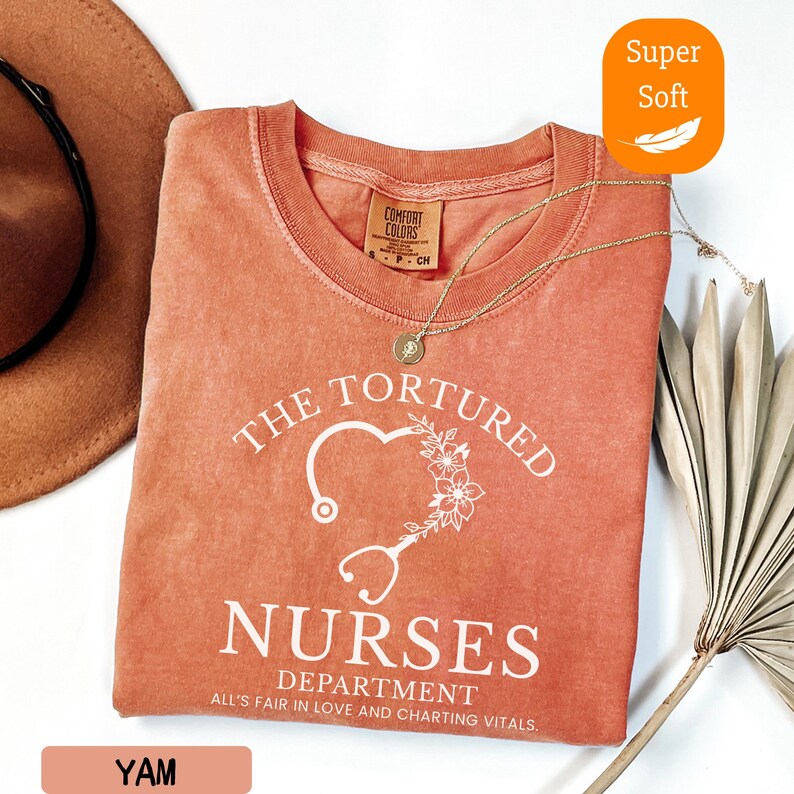 May include: A coral-colored t-shirt with the text "THE TORTURED NURSES DEPARTMENT" and a heart-shaped stethoscope design. The shirt also has the text "ALL'S FAIR IN LOVE AND CHARTING VITALS." A gold necklace rests on the shirt. The shirt is labeled "Super Soft."