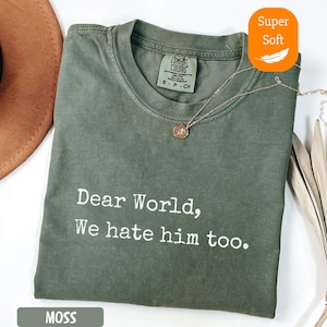 May include: Moss green t-shirt with the text "Dear World, We hate him too." A gold necklace with a pendant is draped on the shirt. The shirt has a tag that says "Comfort Colors".