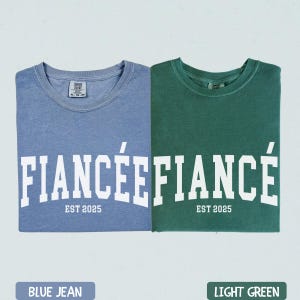 K&ouml;nnte beinhalten: Zwei gefaltete T-Shirts, eines blau und eines gr&uuml;n, mit dem Wort "FIANC&Eacute;E" in Wei&szlig; bedruckt. Darunter steht "EST 2025". Das blaue Shirt ist mit "BLUE JEAN" und das gr&uuml;ne mit "LIGHT GREEN" beschriftet.
