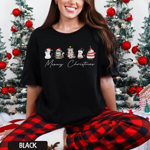 May include: Black t-shirt featuring a festive design with five cartoon cats wearing holiday attire. The text "Meowy Christmas" is written below the cats. The shirt is part of a Christmas-themed collection.