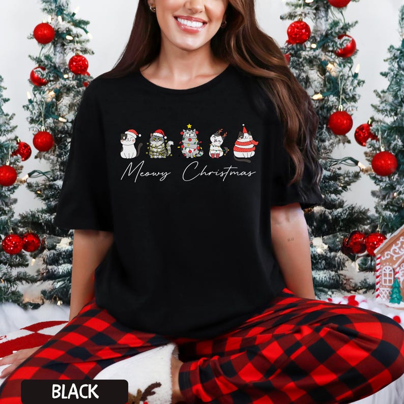 May include: Black t-shirt featuring a festive design with five cartoon cats wearing holiday attire. The text "Meowy Christmas" is written below the cats. The shirt is part of a Christmas-themed collection.