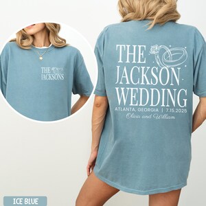 May include: Ice blue t-shirt featuring white text and a wedding ring design. The front displays "The Jacksons". The back reads "The Jackson Wedding, Atlanta, Georgia 7.15.2025, Olivia and William".