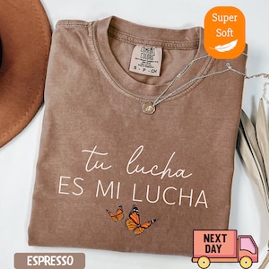 May include: Espresso-colored t-shirt with the text "tu lucha ES MI LUCHA" and two orange butterflies. The shirt has a crew neck and short sleeves. A silver necklace with a pendant is draped on the shirt. The shirt is labeled "Super Soft".