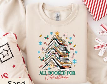 May include: A beige sweatshirt with a Christmas tree made of stacked books. The tree is decorated with lights, candy canes, and holly. The text "All Booked for Christmas" is printed below the tree.