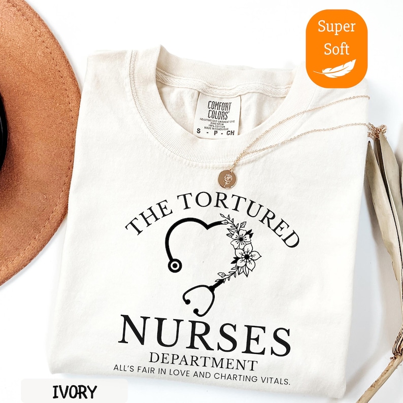 May include: Ivory-colored t-shirt with the text "THE TORTURED NURSES DEPARTMENT" in black. The design includes a heart-shaped stethoscope and floral accents. A gold necklace and a brown hat are partially visible.