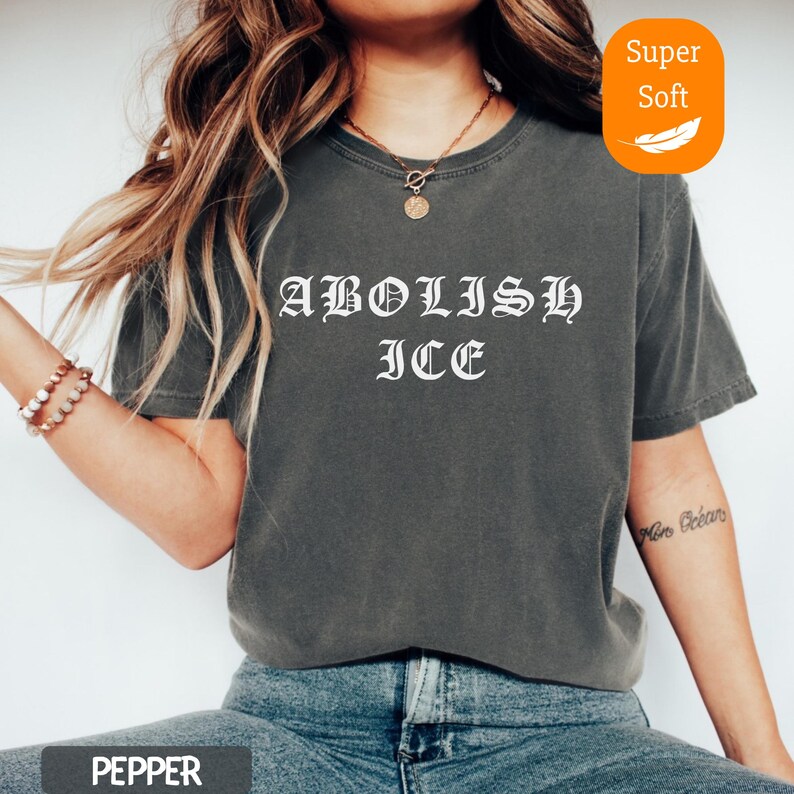 May include: A dark gray t-shirt with the words "ABOLISH ICE" in white gothic lettering. The shirt has a relaxed fit and is made of a soft material. The word "PEPPER" is at the bottom of the image.