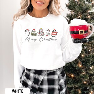May include: White sweatshirt with a graphic of five cats dressed in Christmas attire. The phrase "Meowy Christmas" is written below the cats. A red and white Santa-themed mug is held in the hand.