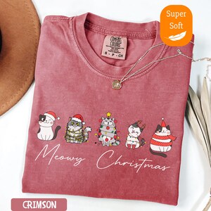 May include: Crimson-colored t-shirt with a crew neck and the words "Meowy Christmas" in white script. The shirt features a graphic of five cats wearing Christmas hats and lights. The shirt is labeled "Super Soft".