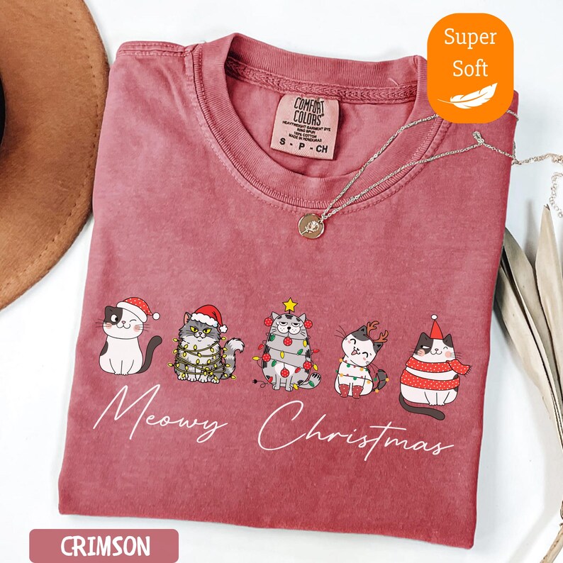 May include: Crimson-colored t-shirt with a crew neck and the words "Meowy Christmas" in white script. The shirt features a graphic of five cats wearing Christmas hats and lights. The shirt is labeled "Super Soft".