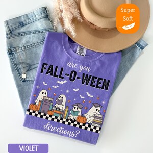 May include: A violet t-shirt with the text "FALL-O-WEEN" and the question "are you directions?" is displayed. The shirt features a graphic of ghosts, pumpkins, books, and bats. The shirt is paired with blue jeans and a tan hat.