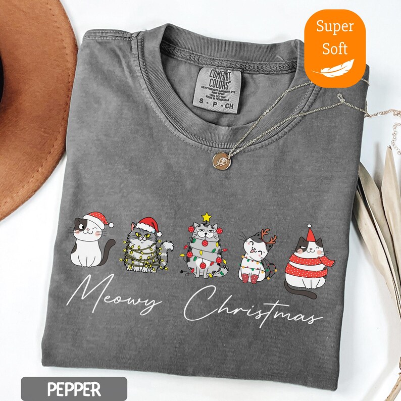 May include: A gray, short-sleeved t-shirt with a festive cat graphic. The design features five cats wearing Christmas hats and lights, with the words "Meowy Christmas" in white script. The shirt is labeled "Super Soft."