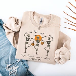 May include: Beige sweatshirt featuring a graphic of three dancing skeletons holding jack-o'-lanterns, with a rising orange sun and black bats. The text "dry bones come alive" and "Ezekiel 37:5" are printed below the graphic.