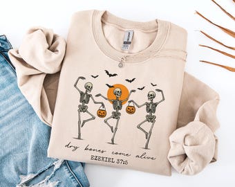 May include: Beige sweatshirt featuring a graphic of three dancing skeletons holding jack-o'-lanterns, with a rising orange sun and black bats. The text "dry bones come alive" and "Ezekiel 37:5" are printed below the graphic.
