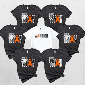 May include: A collection of t-shirts, primarily dark gray, with the text "OLIVIA'S SUPPORT SQUAD" and an orange ribbon graphic. One white t-shirt features the word "WARRIOR" in black. All shirts have a crew neck.