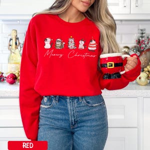 May include: A red sweatshirt featuring a "Meowy Christmas" design with five cartoon cats in festive attire. The cats are wearing hats, scarves, and other holiday accessories. The sweatshirt is paired with blue jeans.