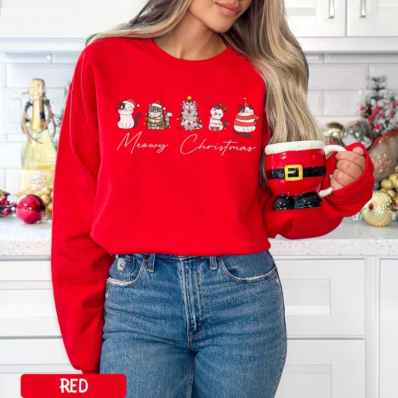 May include: A red sweatshirt featuring a "Meowy Christmas" design with five cartoon cats in festive attire. The cats are wearing hats, scarves, and other holiday accessories. The sweatshirt is paired with blue jeans.