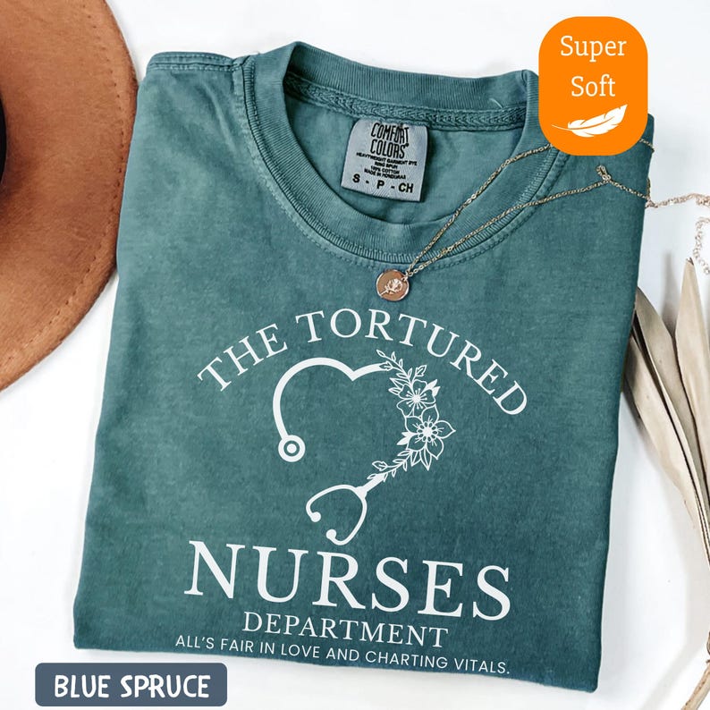 May include: Blue spruce t-shirt with the text "THE TORTURED NURSES DEPARTMENT" and a stethoscope heart design. The shirt also has the text "ALL'S FAIR IN LOVE AND CHARTING VITALS." A gold necklace is draped across the shirt. The shirt is labeled "Super Soft."