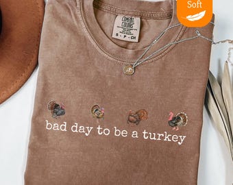 May include: Espresso-colored t-shirt with embroidered turkeys and the phrase "bad day to be a turkey." The shirt has a crew neck and short sleeves. A silver necklace with a pendant is draped on the shirt.