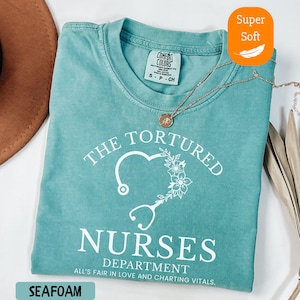 May include: Seafoam green t-shirt with the text "THE TORTURED NURSES DEPARTMENT" and a heart-shaped stethoscope design. The shirt is labeled "Super Soft" and has a Comfort Colors tag. A gold necklace rests on the shirt.