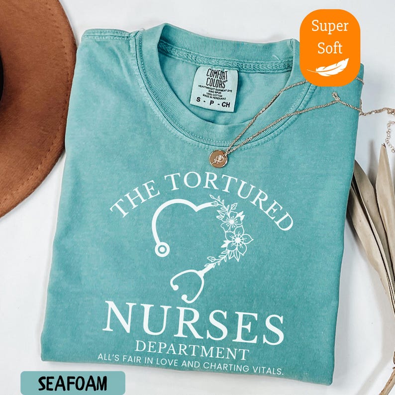May include: Seafoam green t-shirt with the text "THE TORTURED NURSES DEPARTMENT" and a heart-shaped stethoscope design. The shirt is labeled "Super Soft" and has a Comfort Colors tag. A gold necklace rests on the shirt.