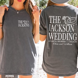 May include: Dark grey t-shirt featuring white text and a wedding ring design. The front displays "The Jacksons." The back reads "The Jackson Wedding" with the date, location, and couple's names.