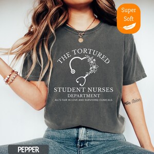 May include: Pepper-colored t-shirt with white text: "THE TORTURED STUDENT NURSES DEPARTMENT." A heart-shaped design with a stethoscope and floral accents is featured. The text "ALL'S FAIR IN LOVE AND SURVIVING CLINICALS" is also present. The shirt has a "Super Soft" label.