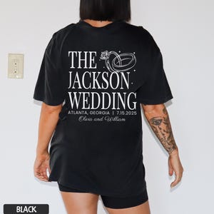 May include: Black t-shirt featuring white text and a ring graphic. The text reads "THE JACKSON WEDDING" with the location and date: Atlanta, Georgia | 7.15.2025. The names Olivia and William are also printed on the shirt.