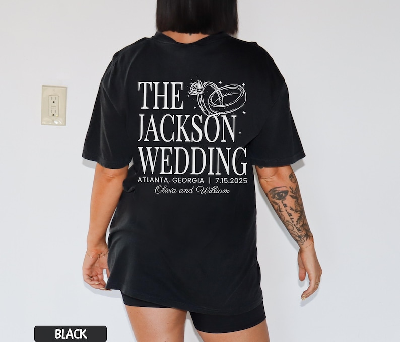 May include: Black t-shirt featuring white text and a ring graphic. The text reads "THE JACKSON WEDDING" with the location and date: Atlanta, Georgia | 7.15.2025. The names Olivia and William are also printed on the shirt.