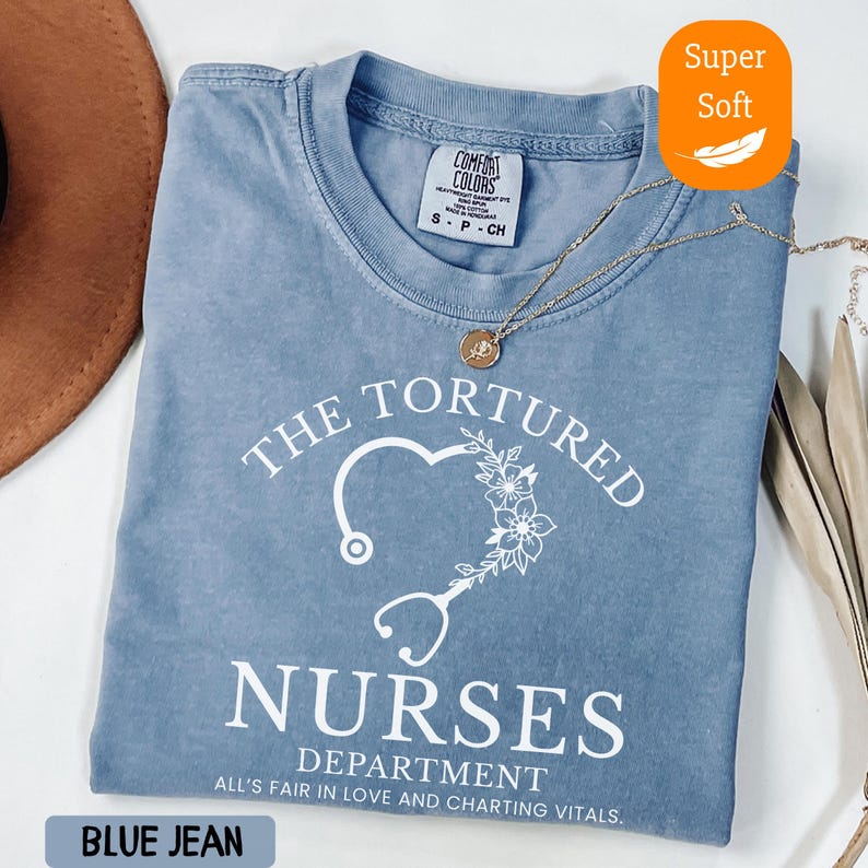 May include: Blue jean colored t-shirt with the text "THE TORTURED NURSES DEPARTMENT" and a heart-shaped stethoscope design. The shirt is labeled "Super Soft" and has a Comfort Colors tag. A gold necklace is draped across the shirt.