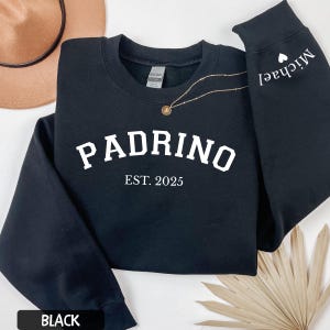 May include: Black sweatshirt with the word "Padrino" and "EST. 2025" printed in white. The sleeve has the name "Michael" printed in white with a small white heart. A gold necklace is draped across the sweatshirt.