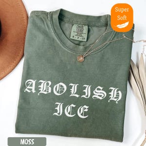 May include: A moss green t-shirt with the words "ABOLISH ICE" in white gothic lettering. The shirt has a crew neck and short sleeves. A gold necklace with a pendant rests on the shirt. The shirt is labeled "Super Soft".