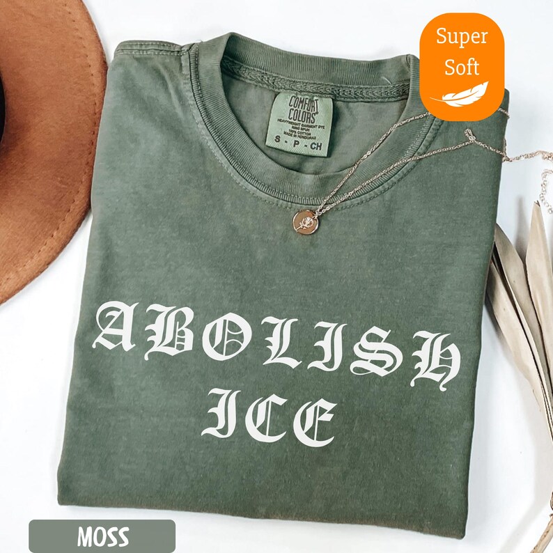 May include: A moss green t-shirt with the words "ABOLISH ICE" in white gothic lettering. The shirt has a crew neck and short sleeves. A gold necklace with a pendant rests on the shirt. The shirt is labeled "Super Soft".
