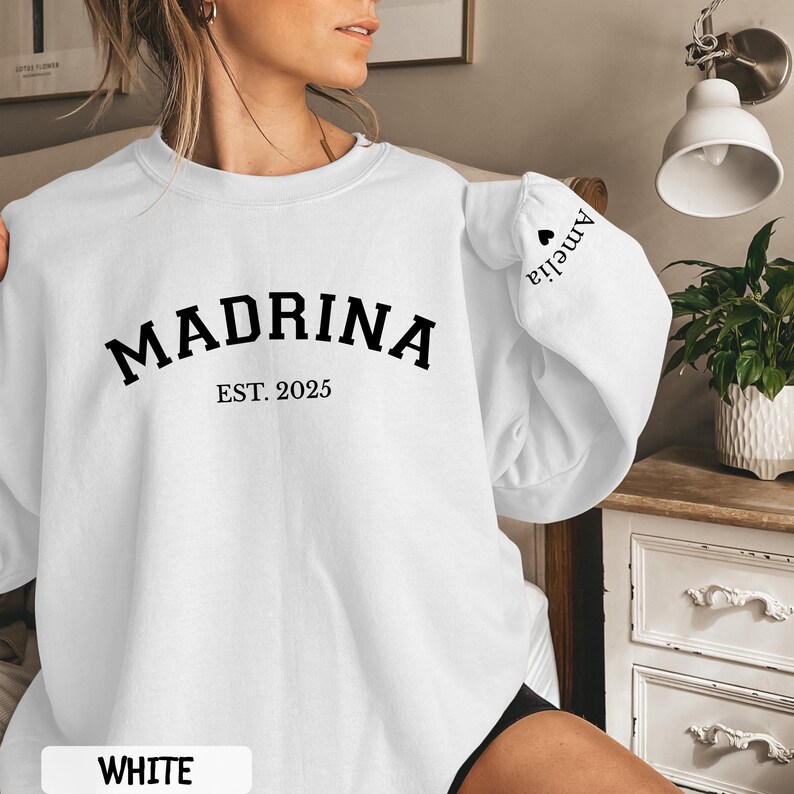 May include: White sweatshirt with the word "MADRINA" in black, with "EST. 2025" underneath. The name "Amelia" is printed on the sleeve. The word "WHITE" is printed at the bottom of the image.
