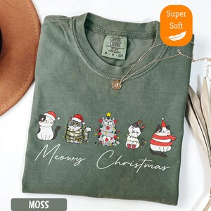 May include: Moss green t-shirt with a "Meowy Christmas" graphic featuring five cartoon cats in festive attire. The shirt is labeled "Super Soft" and has a Comfort Colors tag.