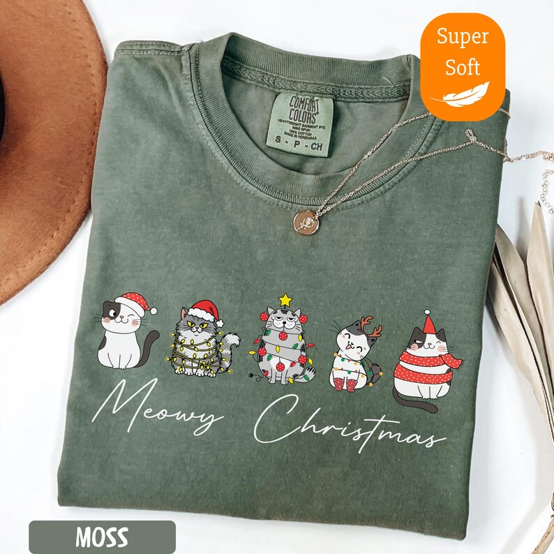 May include: Moss green t-shirt with a "Meowy Christmas" graphic featuring five cartoon cats in festive attire. The shirt is labeled "Super Soft" and has a Comfort Colors tag.