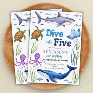 May include: Two invitation cards with a watercolor design featuring sea creatures like sharks, turtles, and whales. The text "Dive into Five" and "Benjamin's 2nd Birthday" are displayed. The cards are on a wooden tray.