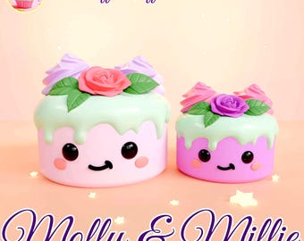 May include: Two pastel-colored cake-shaped figurines with smiling faces, decorated with flowers and frosting. The larger cake is pink and the smaller cake is purple. The text "Molly & Millie" is written below the cakes.