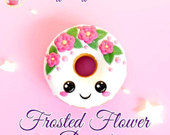 May include: A white frosted donut with pink flowers and green leaves around the top. The donut has a happy face with black eyes and a smile. The donut is on a pink background with the text "Puff Puff Pastries" and "Frosted Flower Donut" in purple.