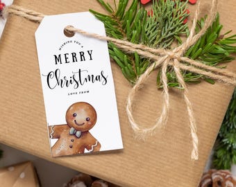 May include: A white gift tag with the text "Wishing you a Merry Christmas Love from" and a gingerbread man illustration. The tag is tied with twine to a brown gift box wrapped with brown paper and decorated with a green pine sprig and a red snowflake.